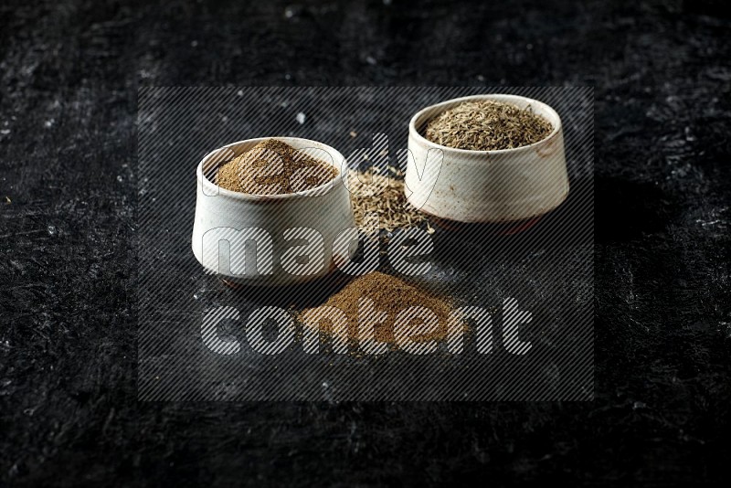 2 beige bowls, one full of cumin powder and the other full of seeds on a textured black flooring