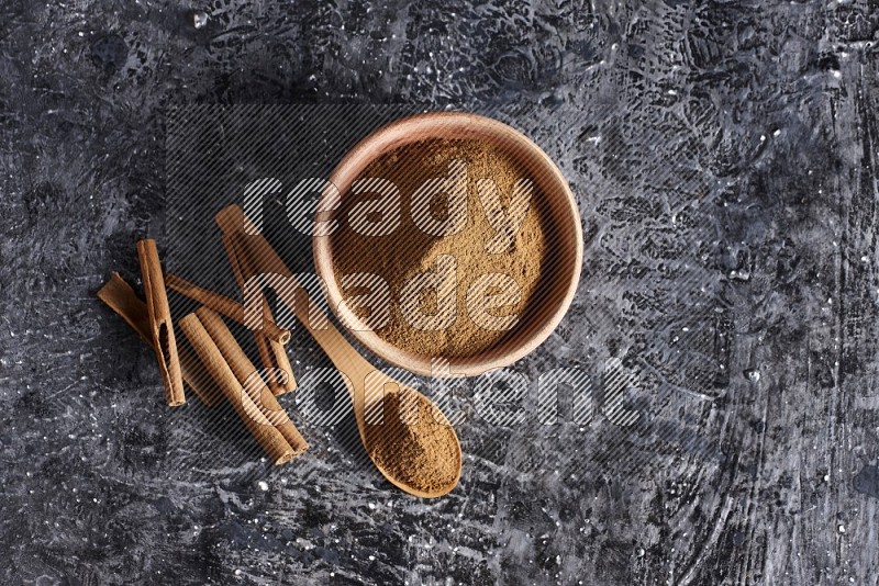 wooden bowl full of cinnamon powder and a wooden spoon full of it with cinnamon sticks on a textured black background