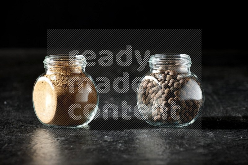 2 glass spice jars full of allspice powder and whole balls on a textured black flooring