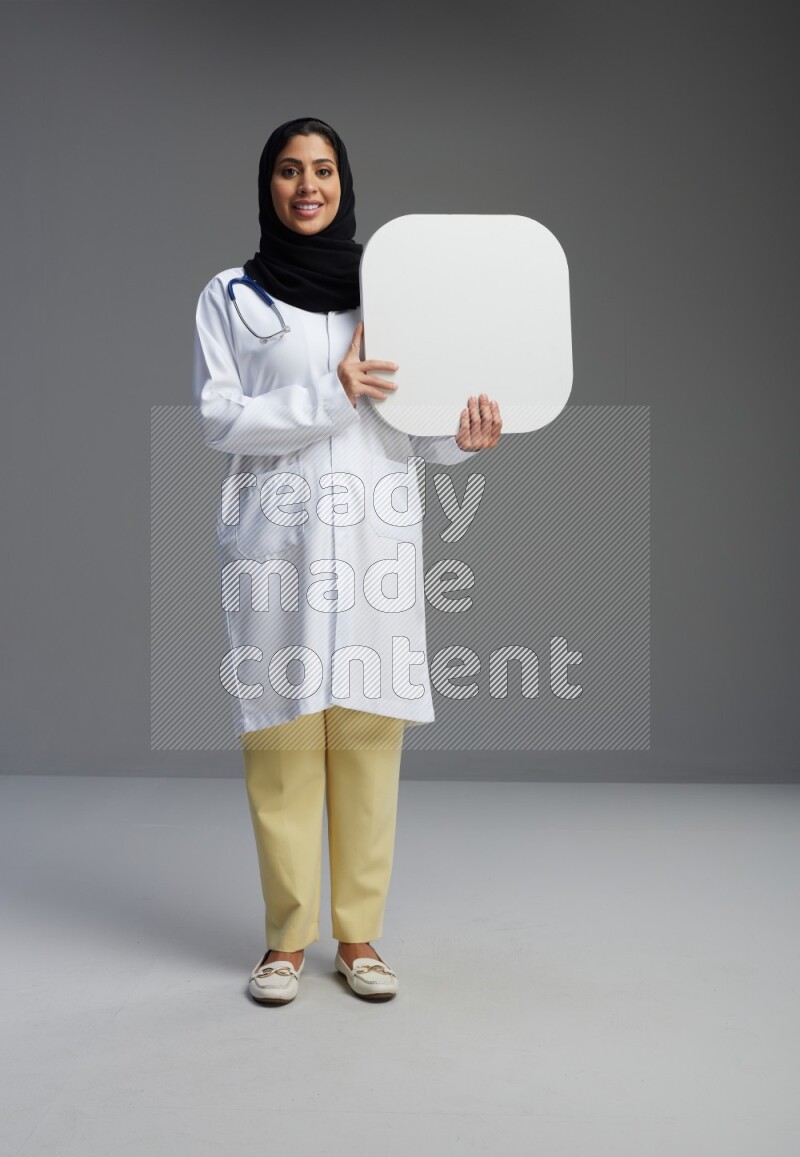 Saudi woman wearing lab coat with stethoscope standing holding social media sign on Gray background