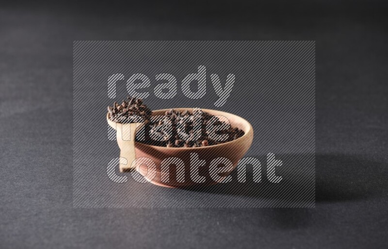 A wooden bowl and a wooden spoon full of cloves on a black flooring