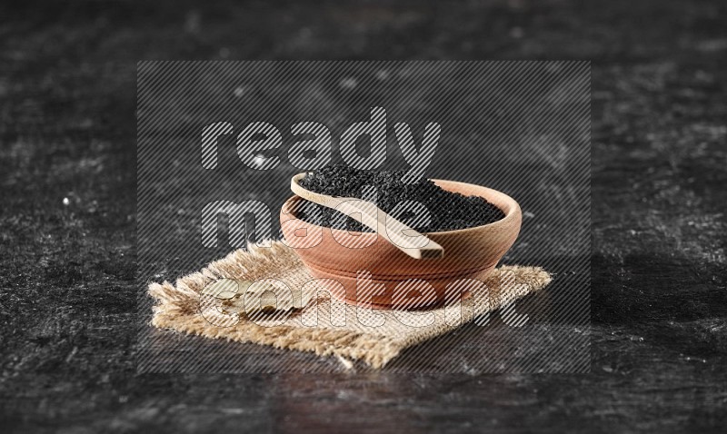 وعاء خشبي مع ملعقة خشبية ممتلئان ببذور حبة البركة علي قطعة من القماش علي خلفية سوداء