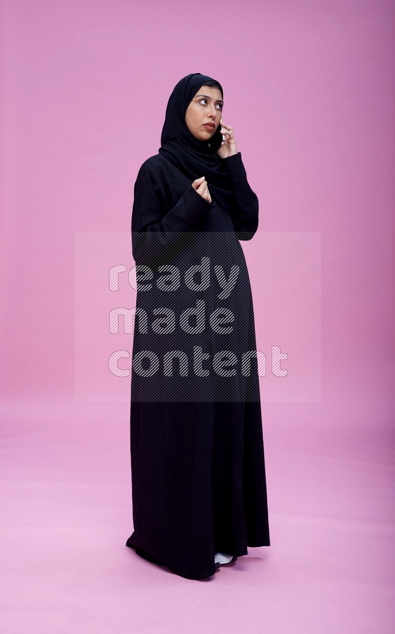 Saudi woman wearing Abaya standing talking on phone on pink background