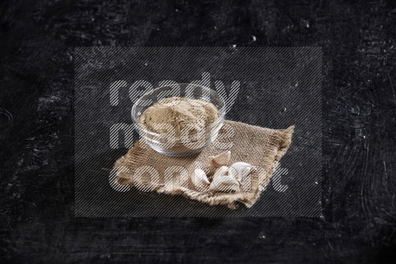 A glass bowl full of garlic powder on burlap fabric with garlic cloves on a textured black flooring in different angles