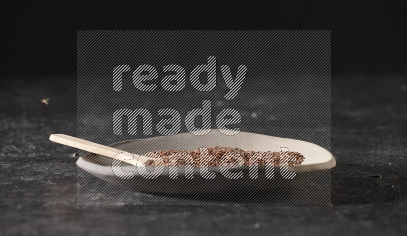 طبق فخار متعدد الألوان ممتلئ بحبوب بذر الكتان وملعقة خشبية ممتلئة بالبذور على أرضية سوداء