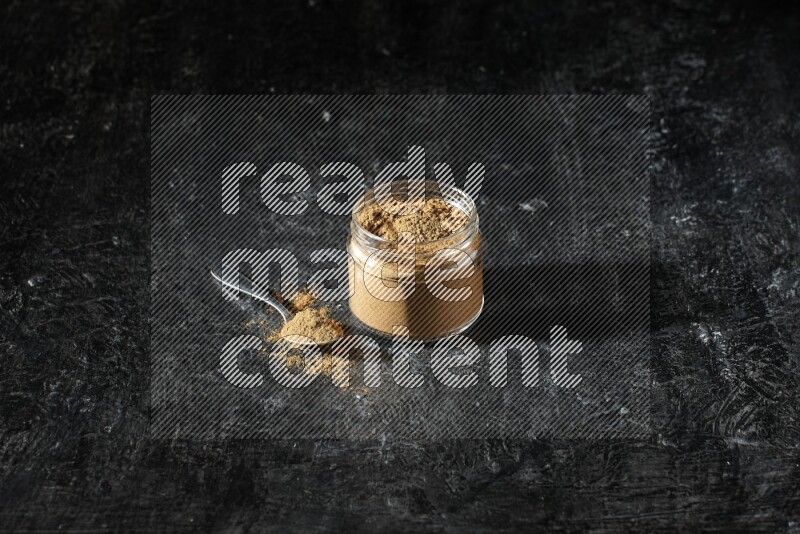 A glass jar and a metal spoon full of allspice powder on a textured black flooring