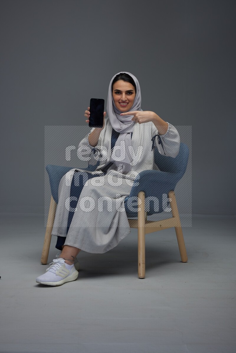 A Saudi woman wearing a light gray Abaya and head scarf sitting on a dark grey chair and showing the phone's screen eye level on a grey background