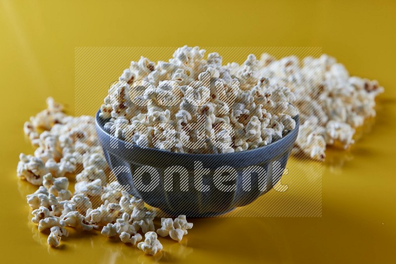 A blue pottery bowl full of popcorn with popcorn beside it on a yellow background in different angles