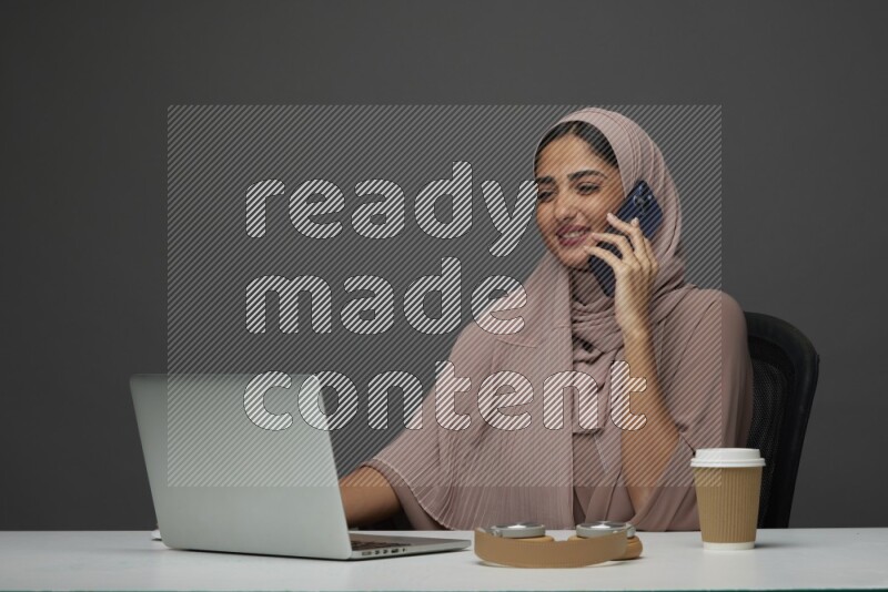 A Saudi woman Setting on her desk
 calling  on a Gray Background wearing Brown Abaya with Hijab