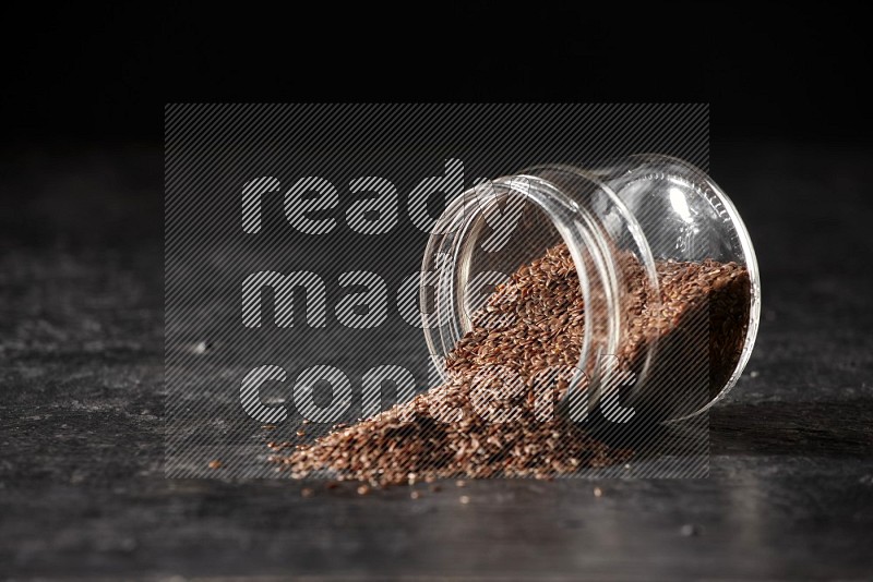A glass jar full of flaxseeds flipped and seeds spread out on a textured black flooring