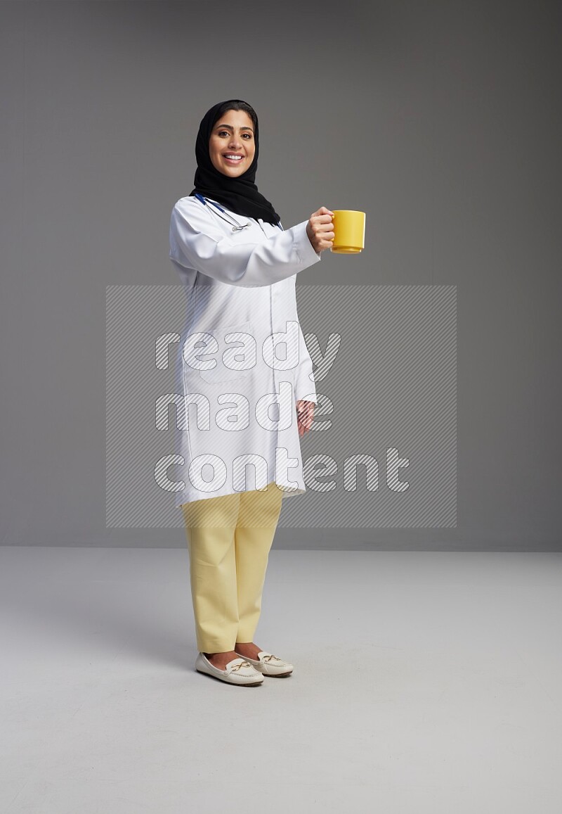Saudi woman wearing lab coat with stethoscope standing holding mug on Gray background