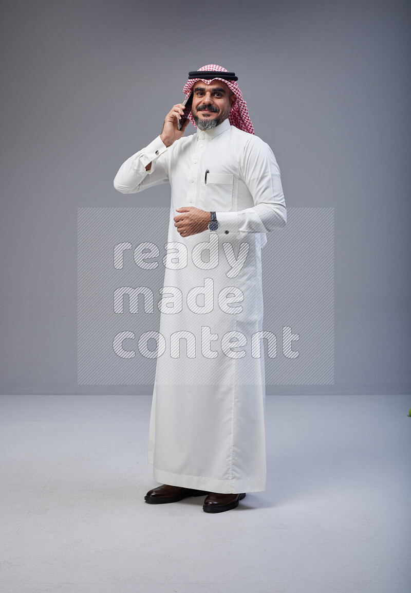 Saudi man Wearing Thob and red Shomag standing talking on phone on Gray background