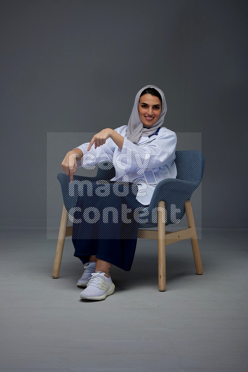 A doctor wearing a light gray head scarf setting on a dark grey chair and using a phone eye level on a grey background