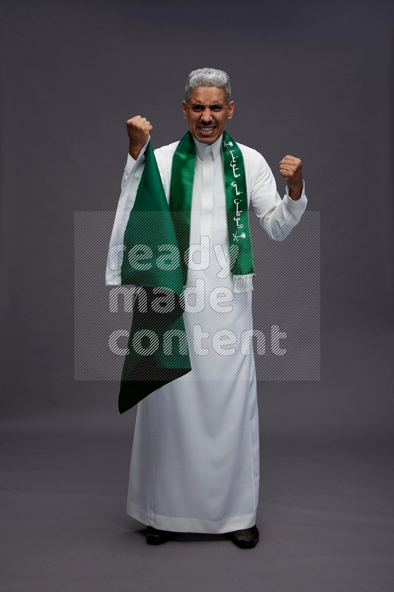 Saudi man wearing thob standing holding Saudi flag on gray background