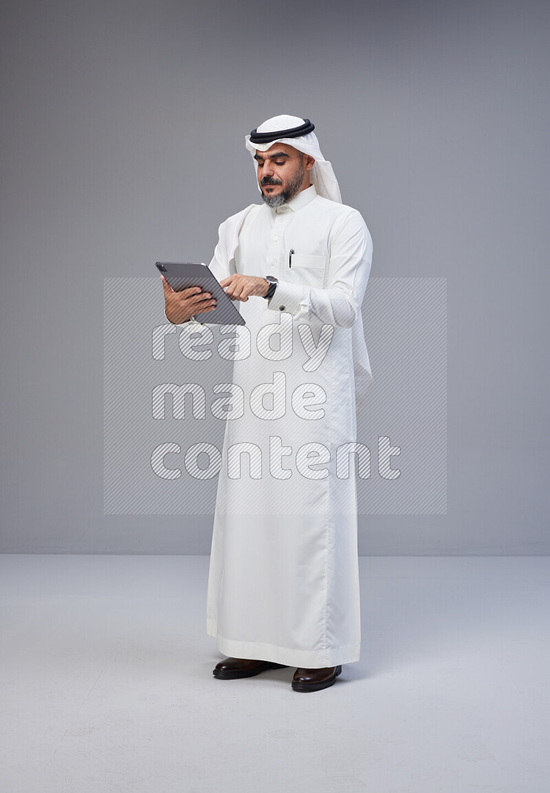 Saudi man Wearing Thob and white Shomag standing working on tablet on Gray background