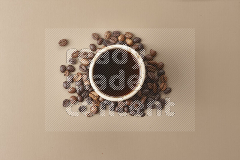 A beige pottery cup of coffee surrounded by roasted coffee beans on beige background