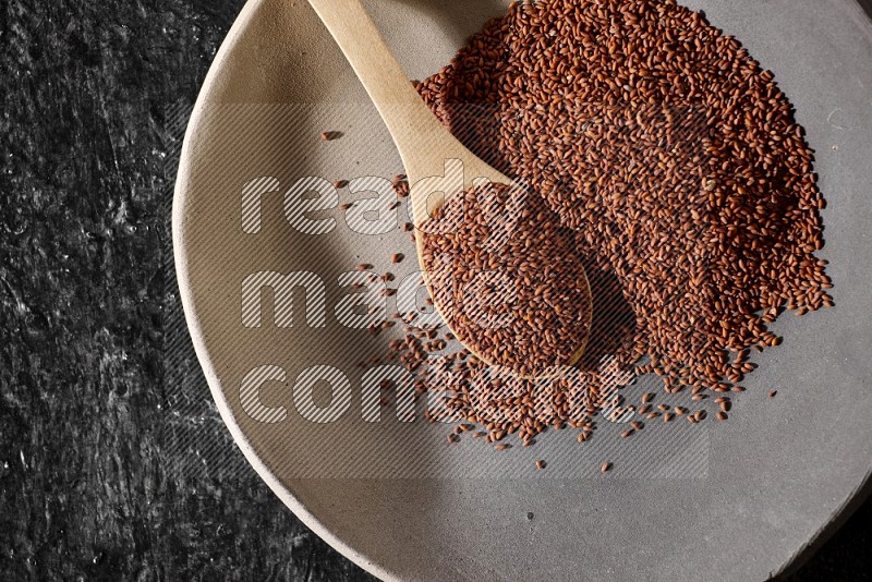 A multicolored pottery plate full of garden cress seeds and wooden spoon full of seeds on a textured black flooring