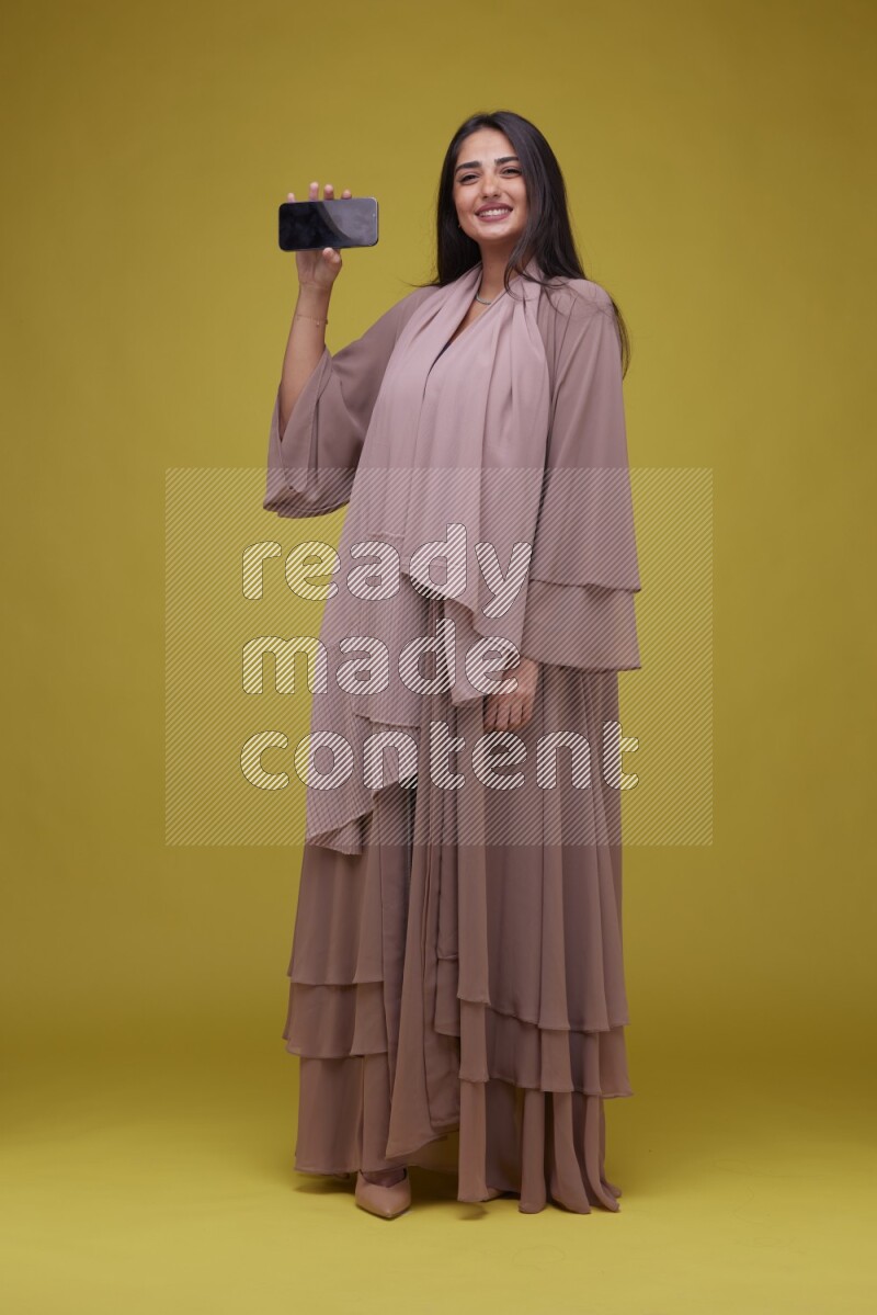 A woman Showing SmartPhone Screen on a Yellow Background wearing Brown Abaya