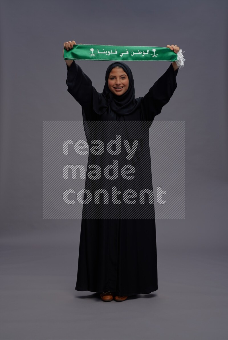Saudi woman wearing Abaya standing holding Saudi flag on gray background