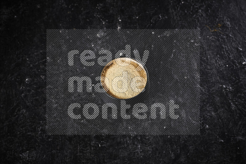 A black pottery bowl full of ground ginger powder on black background