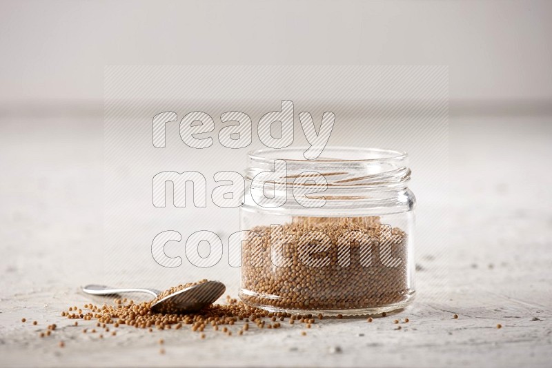 A glass jar and a metal spoon full of mustard seeds on a textured white flooring