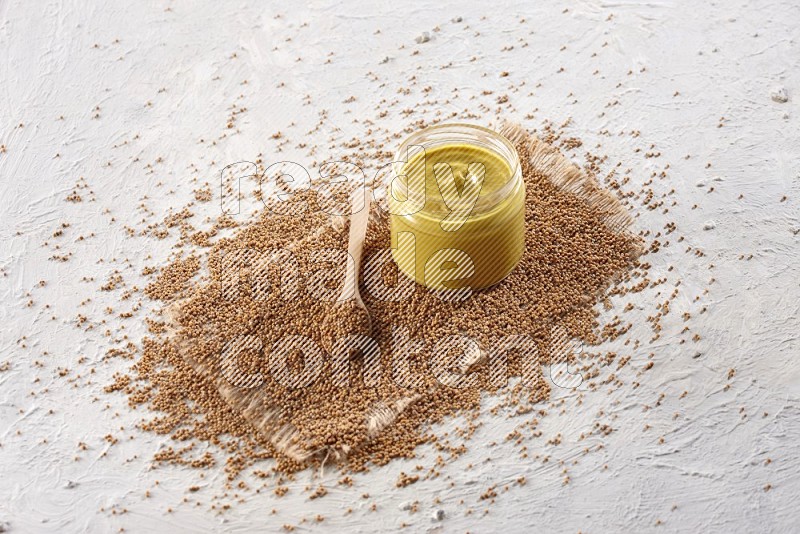 A glass jar full of mustard paste set on a burlap piece and a wooden spoon full of mustard seeds on a textured white flooring in different angles