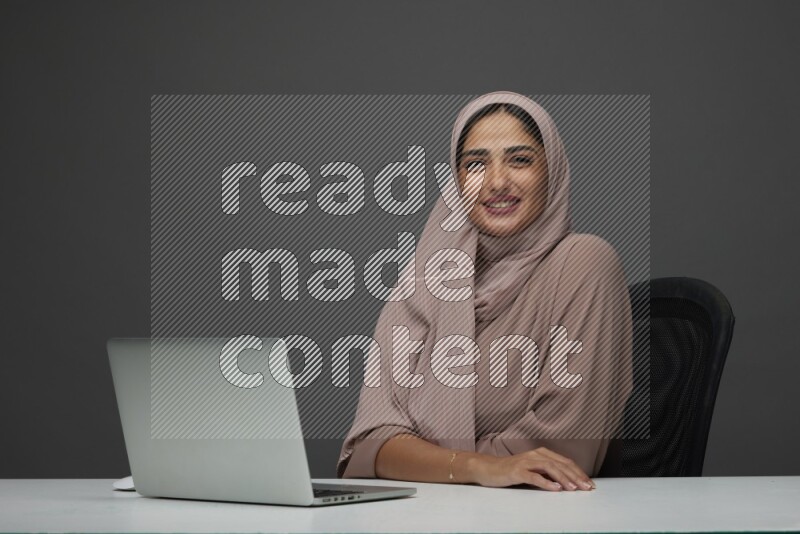 A Saudi woman Setting on her desk on a Gray Background wearing Brown Abaya with Hijab
