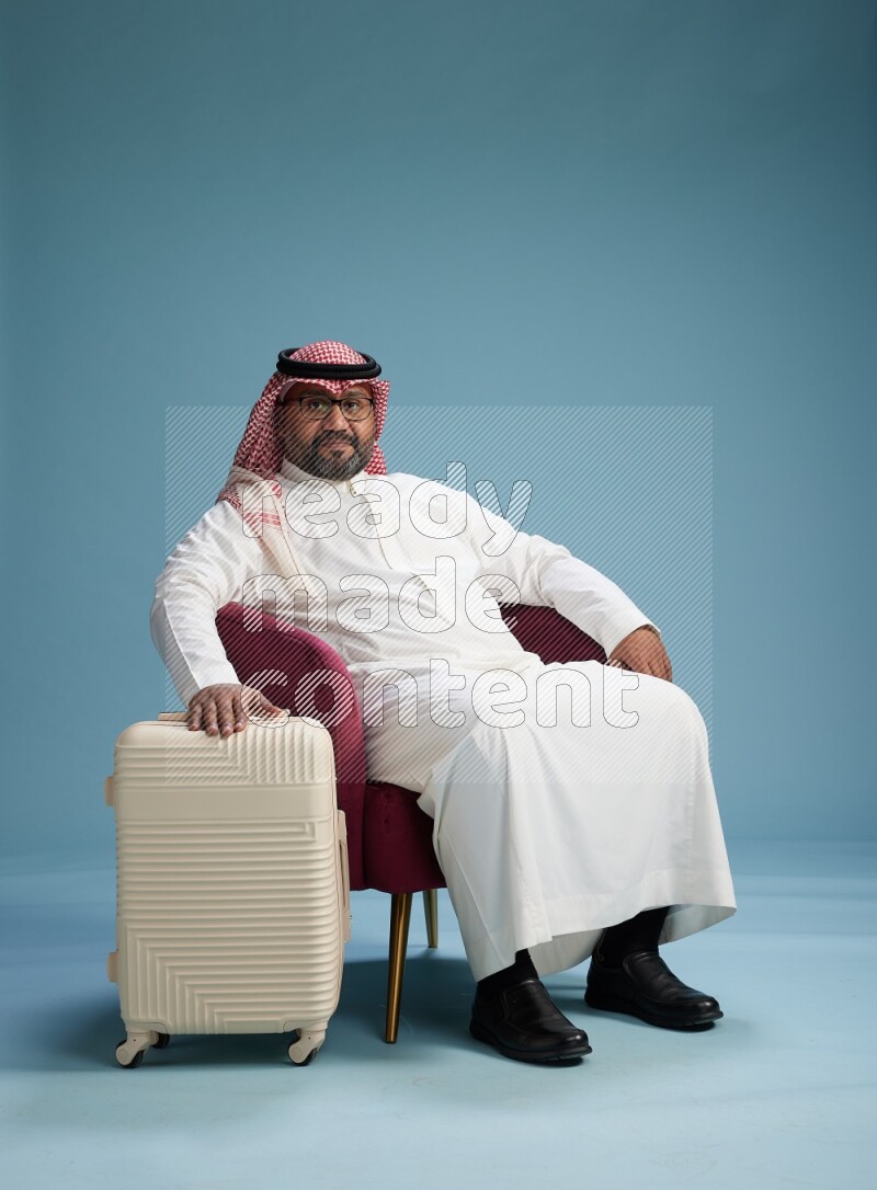 Saudi Man with shimag sitting on chair with a travel luggage on blue background