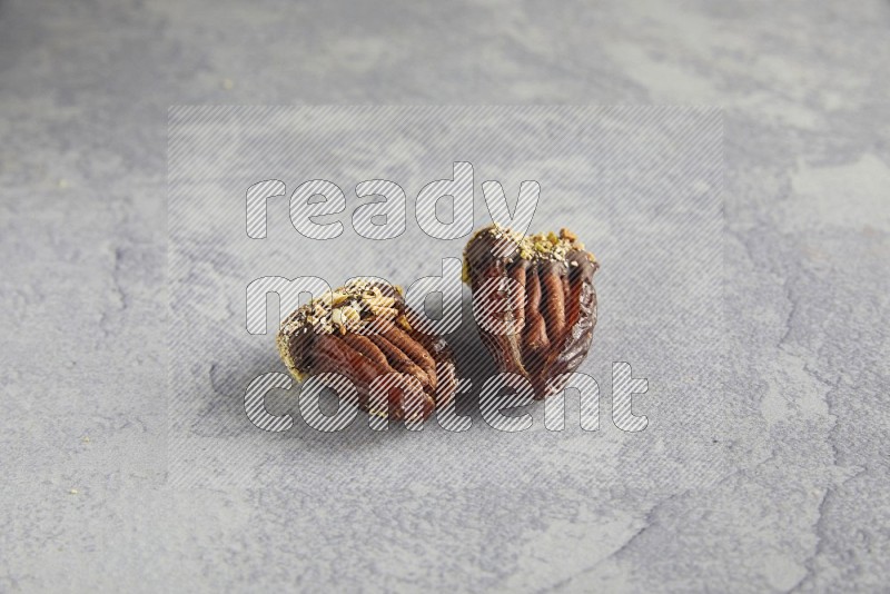 Two pecan stuffed dates covered with Dark chocolate and chopped pistachios on alight grey background
