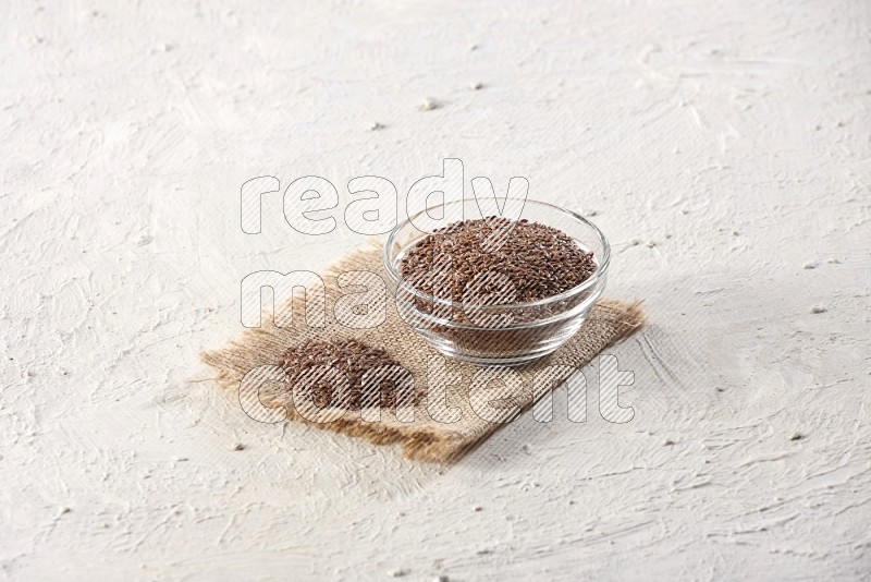 A glass bowl full of flax seeds and a bunch of seeds on burlap fabric on a textured white flooring