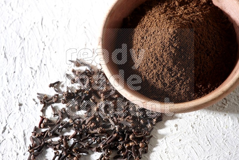 A wooden bowl full of cloves powder with whole cloves beside it on a textured white flooring