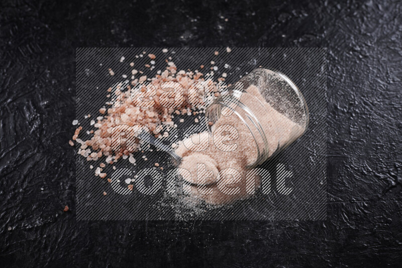 A glass jar full of fine himalayan salt with some himalayan crystals beside it on a black background