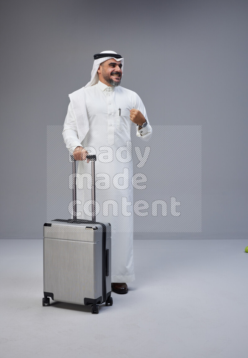 Saudi man wearing Thob and white Shomag standing holding Travel bag on Gray background