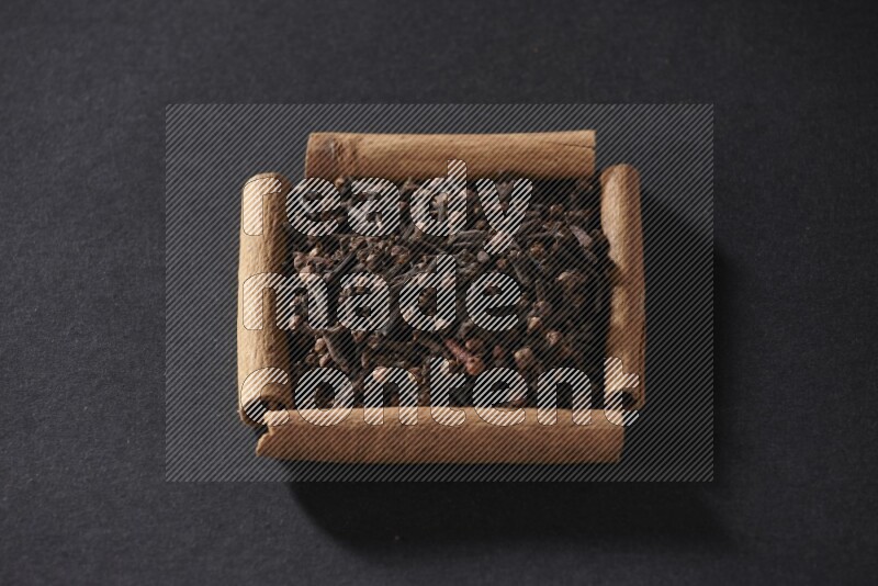 أربعة من أعواد القرفة مكونة شكل مربع بداخلها حبوب قرنفل علي خلفية سوداء