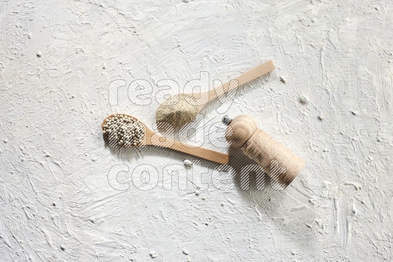 2 wooden spoons one full of white pepper powder and the other with pepper beads and a wooden pepper grinder on textured white flooring