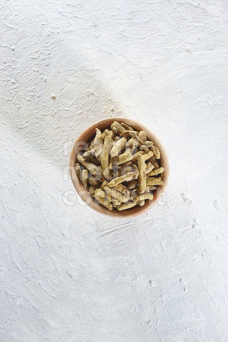 A wooden bowl full of dried turmeric whole fingers on a textured white flooring