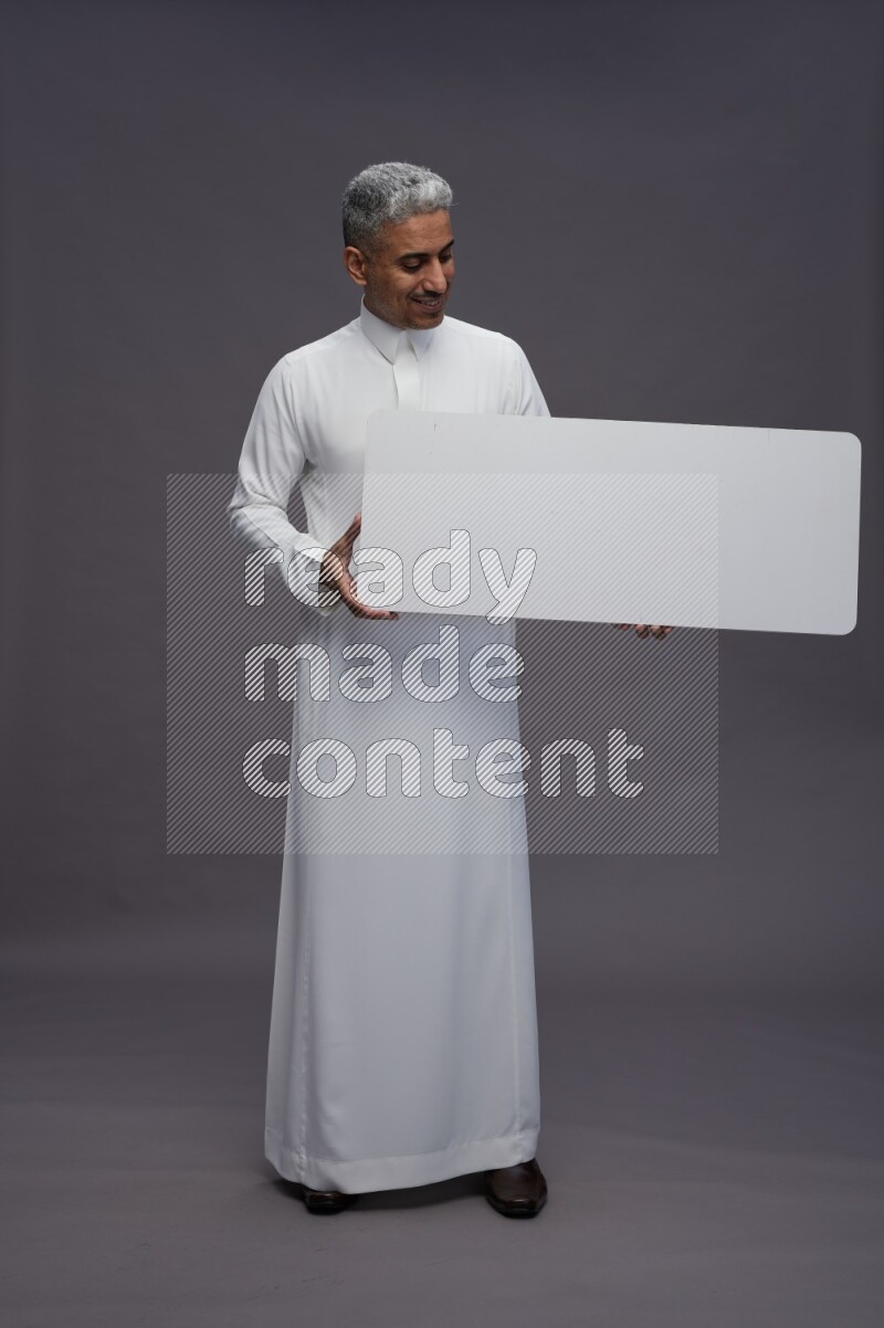 Saudi man wearing thob standing holding board on gray background
