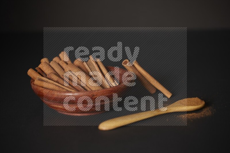 Cinnamon sticks in wooden bowl and cinnamon powder in a wooden spoon on black background