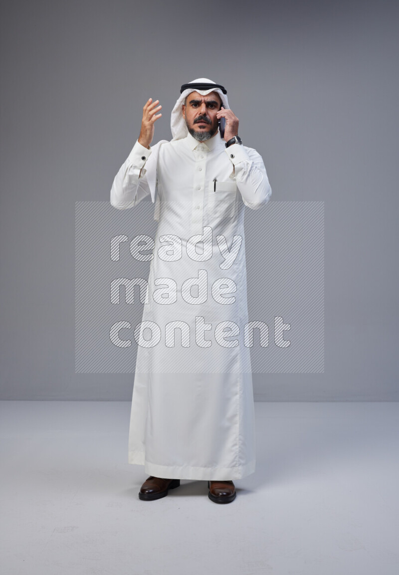 Saudi man Wearing Thob and white Shomag standing talking on phone on Gray background