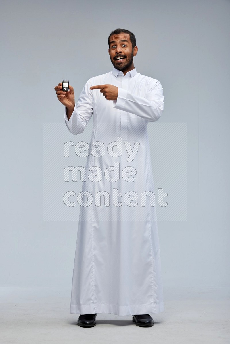 Saudi man wearing thob and shomag standing holding car key on gray background