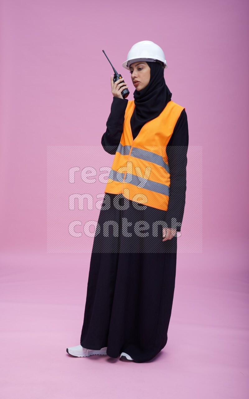 Saudi woman wearing Abaya with engineer vest and helmet standing holding walkie-talkie on pink background