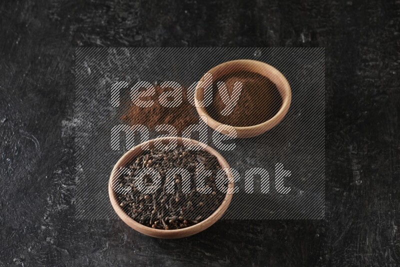 2 wooden bowls full of cloves powder and whole cloves on a textured black flooring