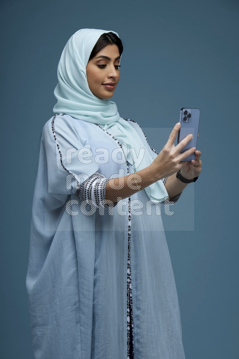 A Saudi woman Taking a Selfie on a blue background wearing a blue Abaya with hijab