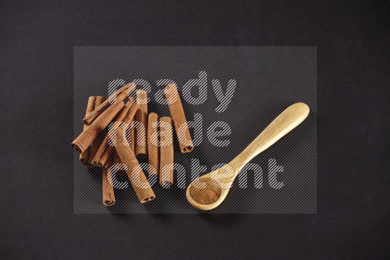 Cinnamon sticks stacked beside a wooden spoon full of cinnamon powder on black background