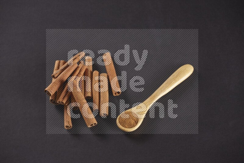 Cinnamon sticks stacked beside a wooden spoon full of cinnamon powder on black background