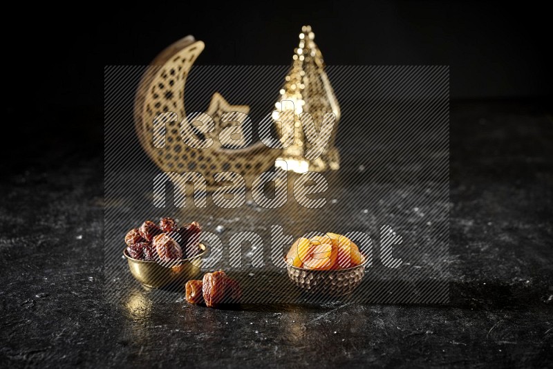 Dates in a metal bowl with dried apricot beside golden lanterns in a dark setup