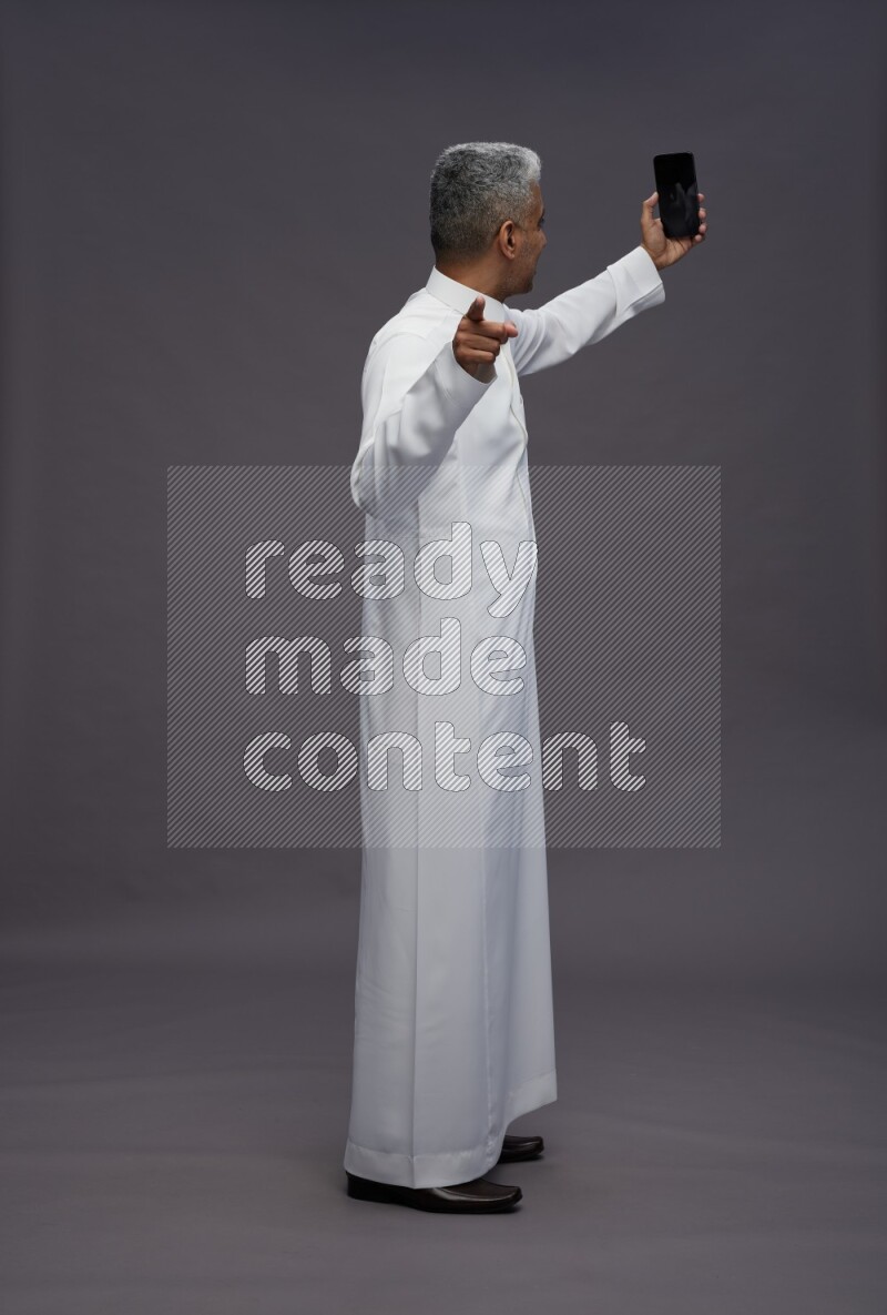 Saudi man wearing thob standing taking selfie on gray background