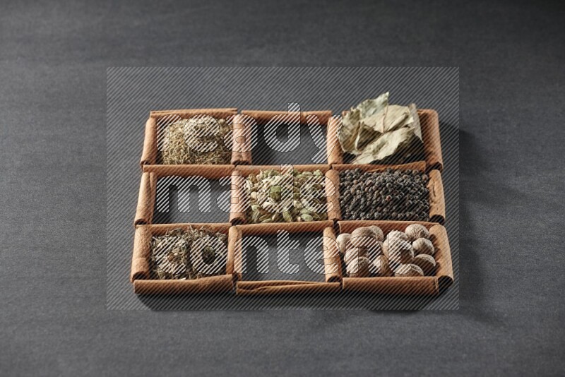9 squares of cinnamon sticks full of cardamom in the middle surrounded by nutmeg, black pepper, bay laurel leaves, allspice, cumin, white pepper, dried basil and salt on black flooring