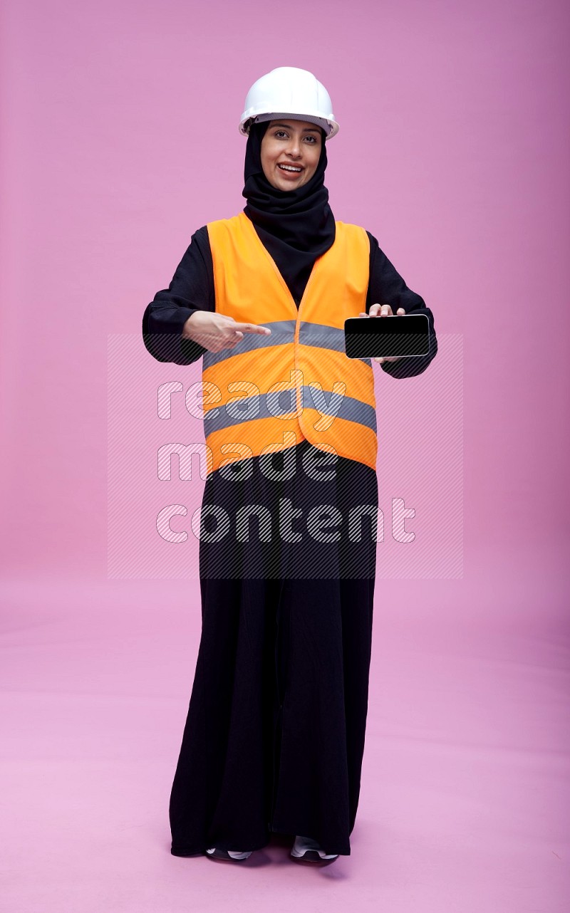 Saudi woman wearing Abaya with engineer vest and helmet standing showing phone to camera on pink background