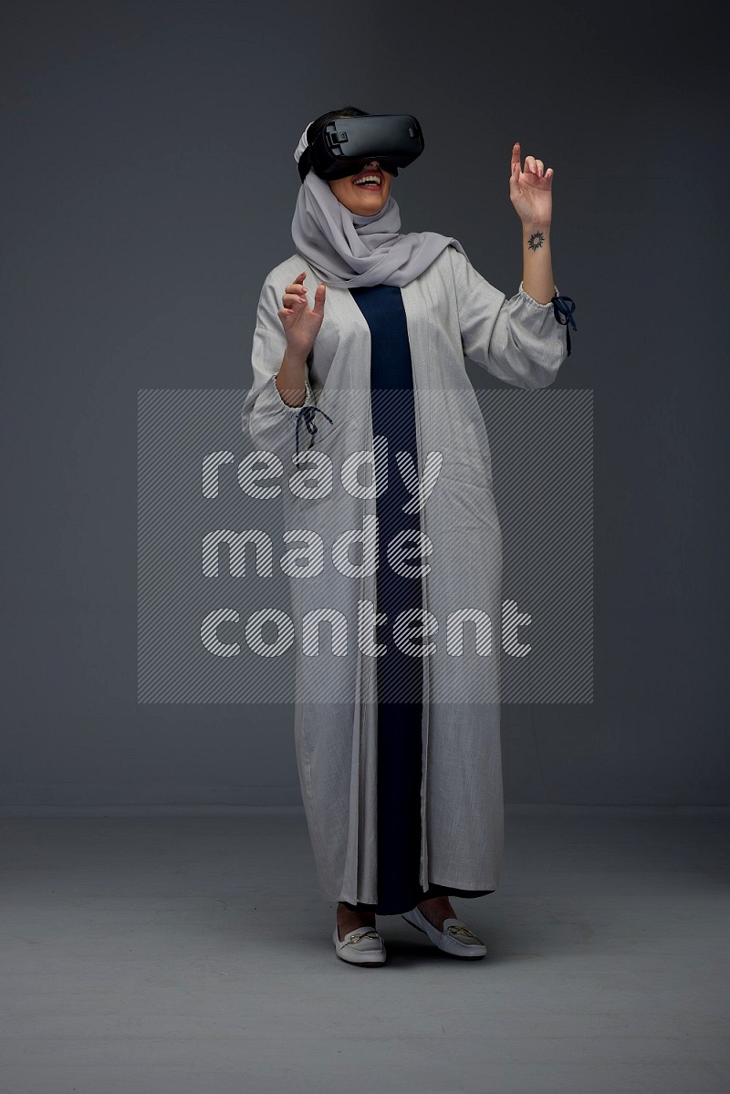 A Saudi woman wearing a light gray Abaya and head scarf standing and wearing VR eye level on a grey background